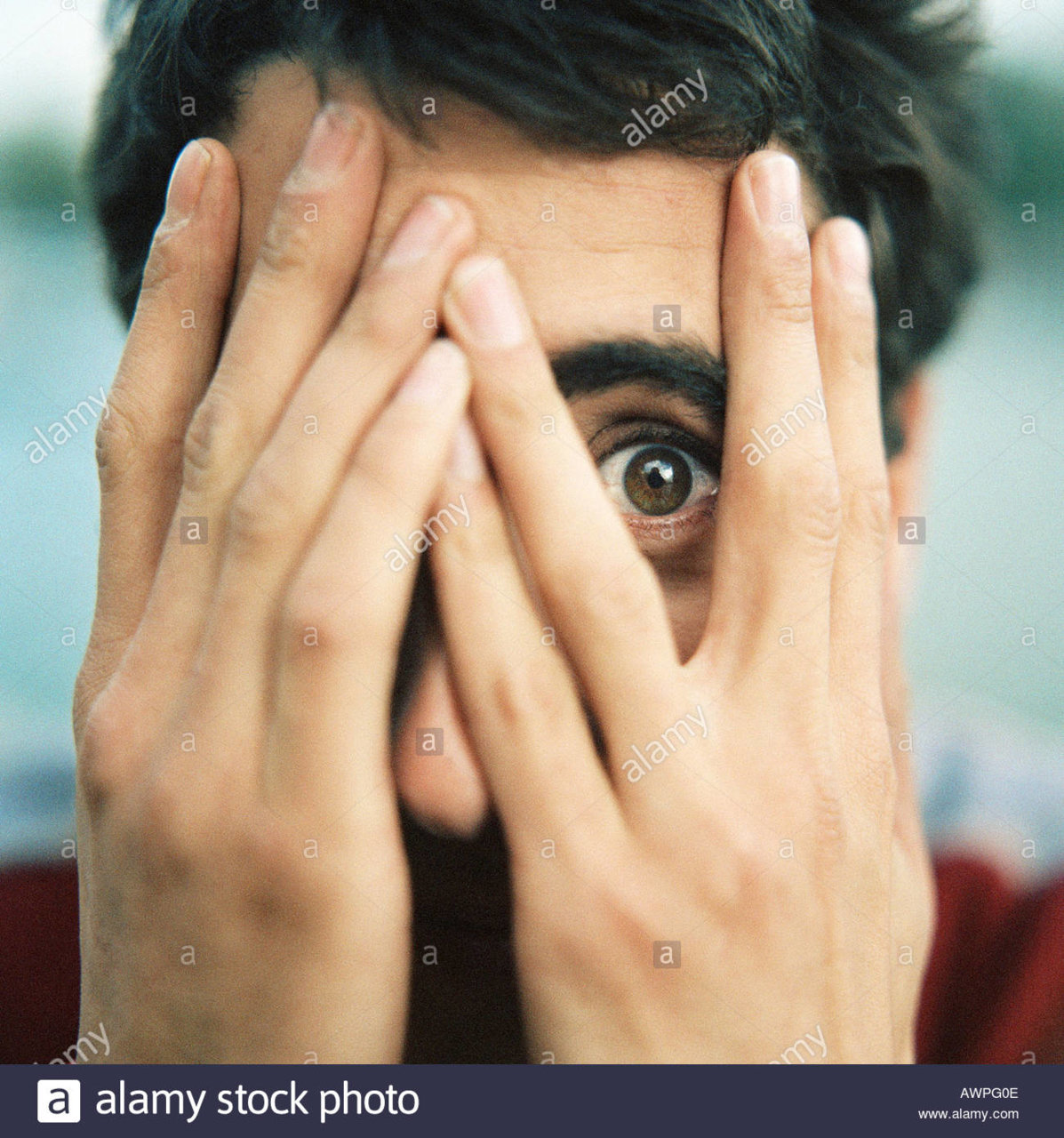 young-man-with-hands-over-face-looking-through-fingers-with-one-eye-AWPG0E.jpg